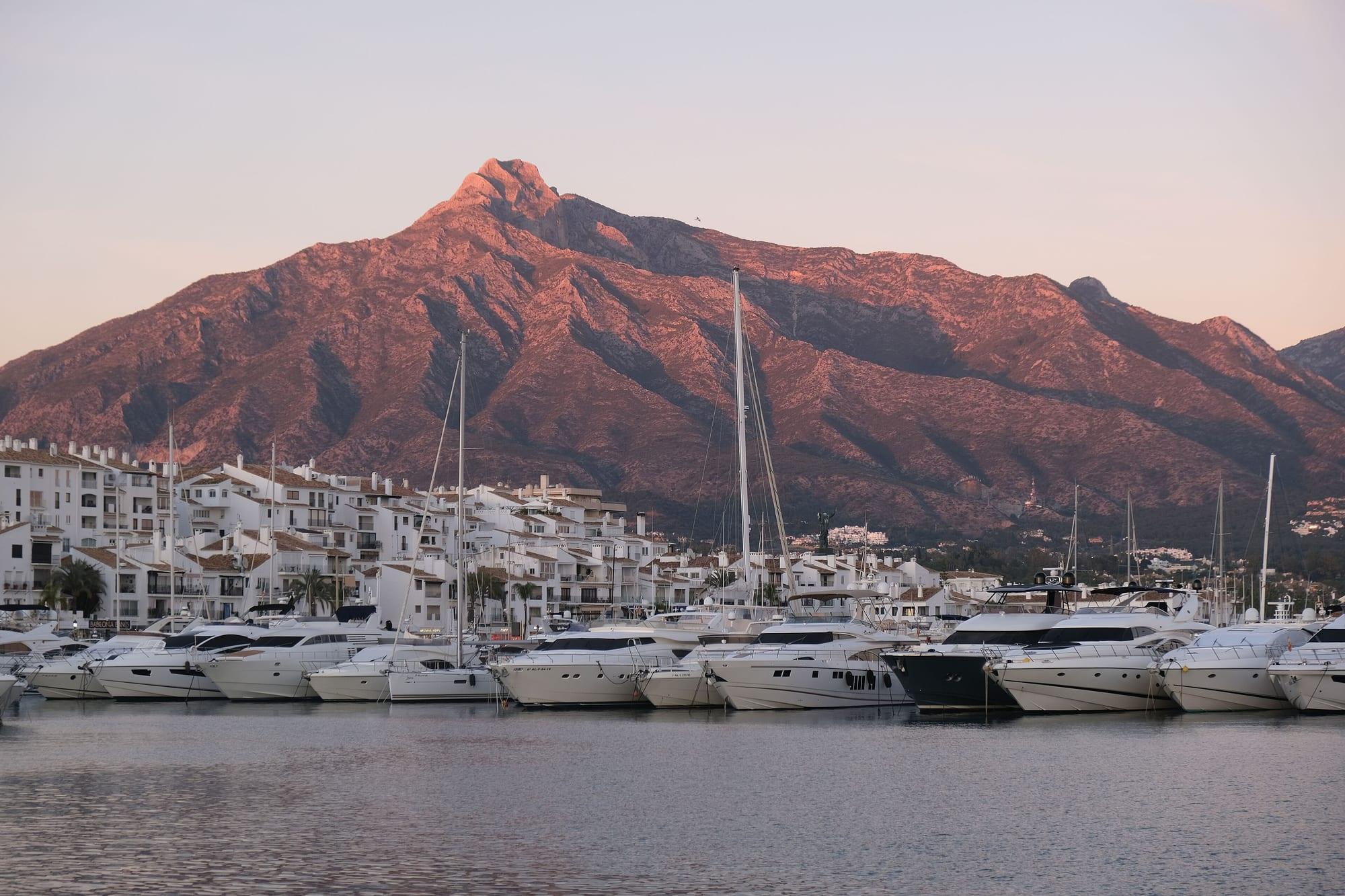 Yachts moored in the Puerto Banús marina at dusk, with white apartment blocks and La Concha peak of Sierra Blanca glowing pink behind