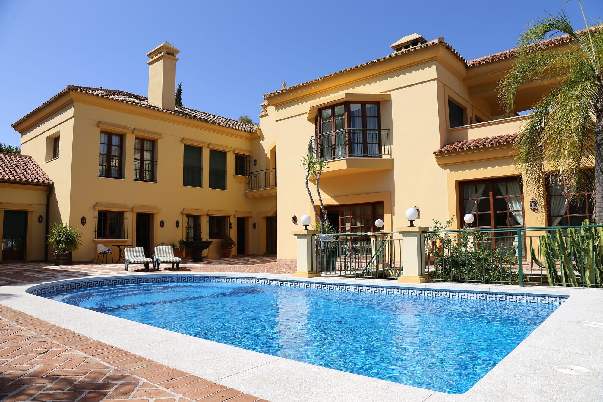 Two-storey Andalusian-style villa in ochre with terracotta roof tiles, palm trees and a blue swimming pool on a tiled terrace