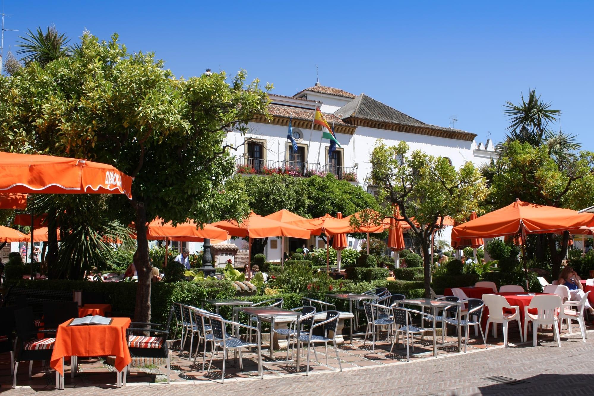 Orange café parasols and terrace tables under orange trees in the Plaza de los Naranjos, with Marbella's whitewashed town hall and its flags behind