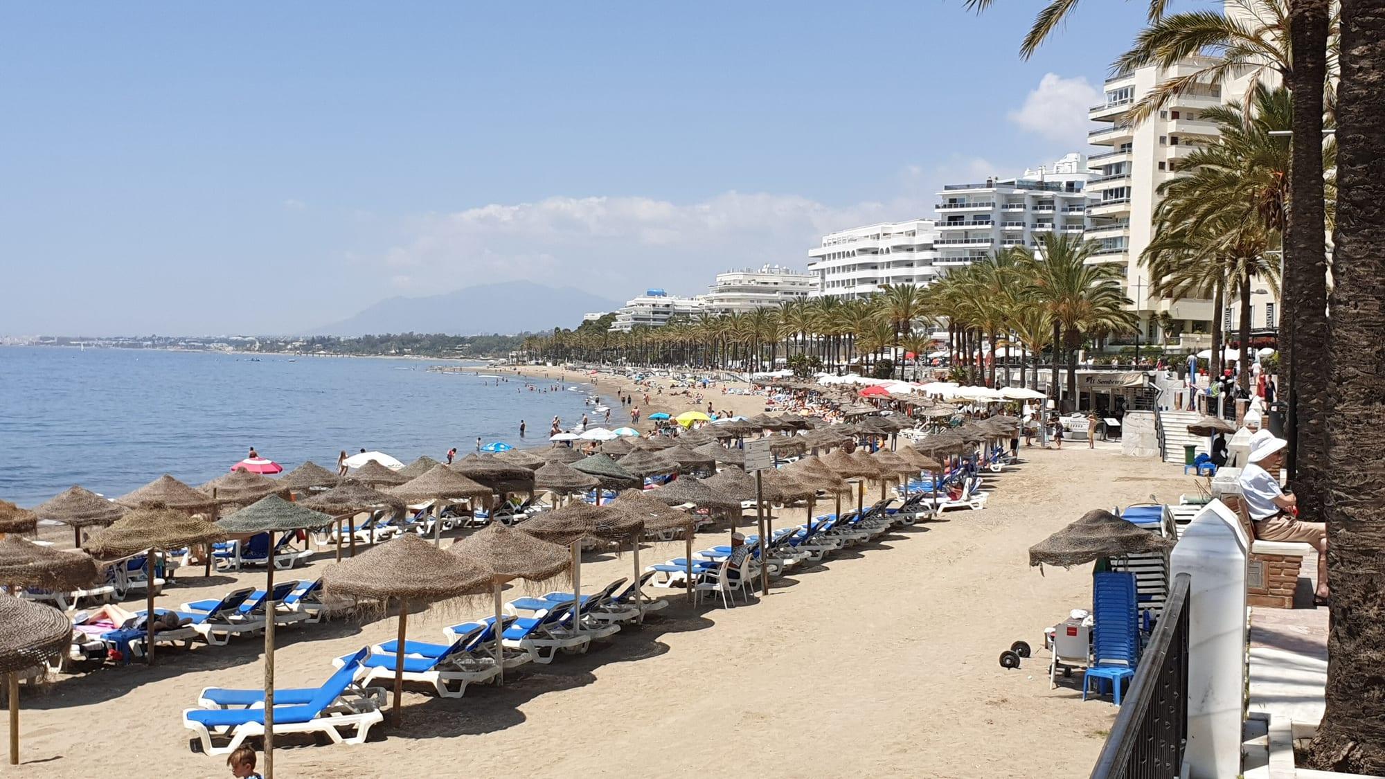 Playa de la Fontanilla in central Marbella: rows of straw parasols and blue sunloungers along the sand, palm-lined promenade and seafront apartment blocks